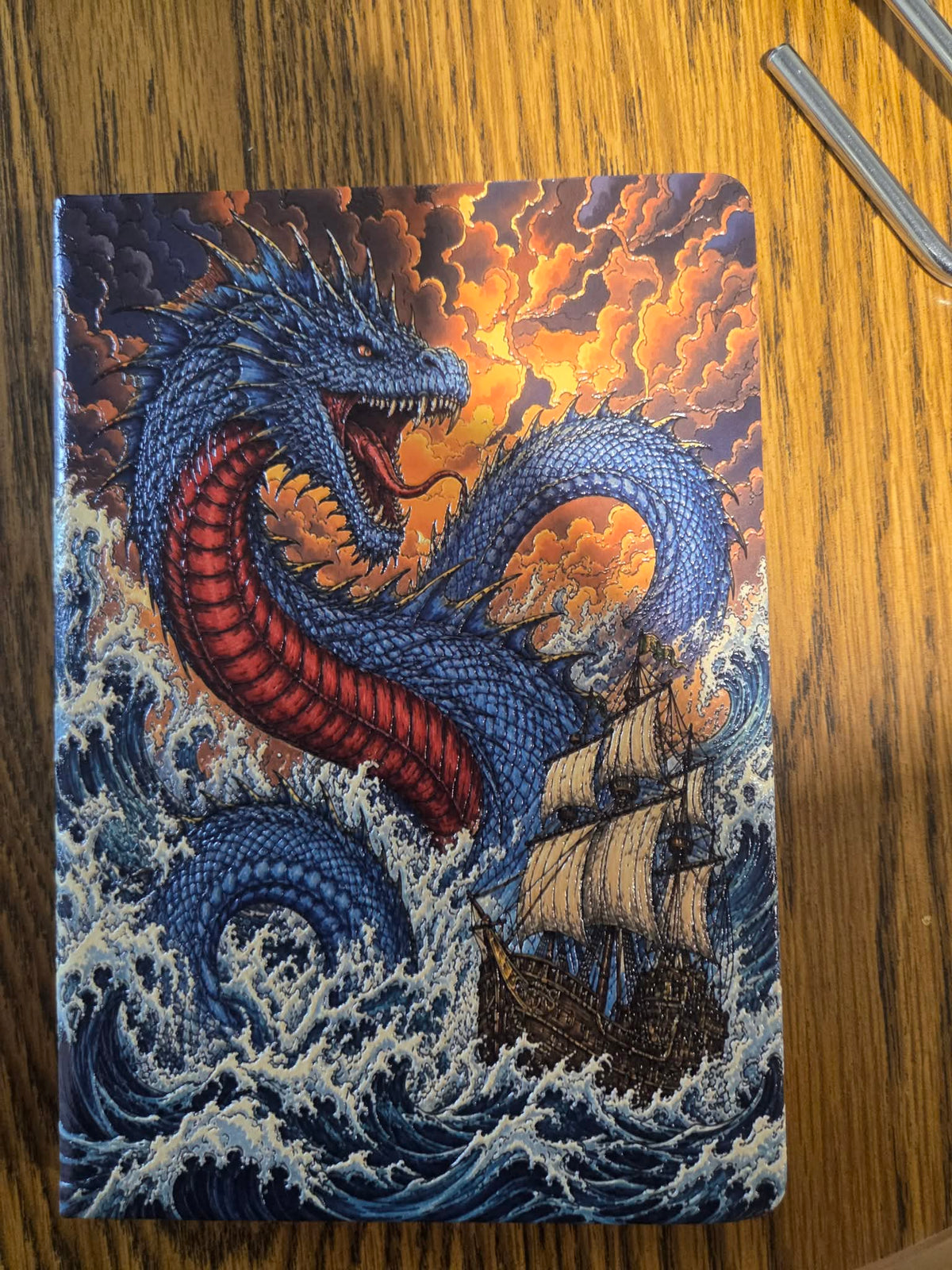 Sea serpent artwork on a hardcover notebook beside a sailing ship in stormy waves